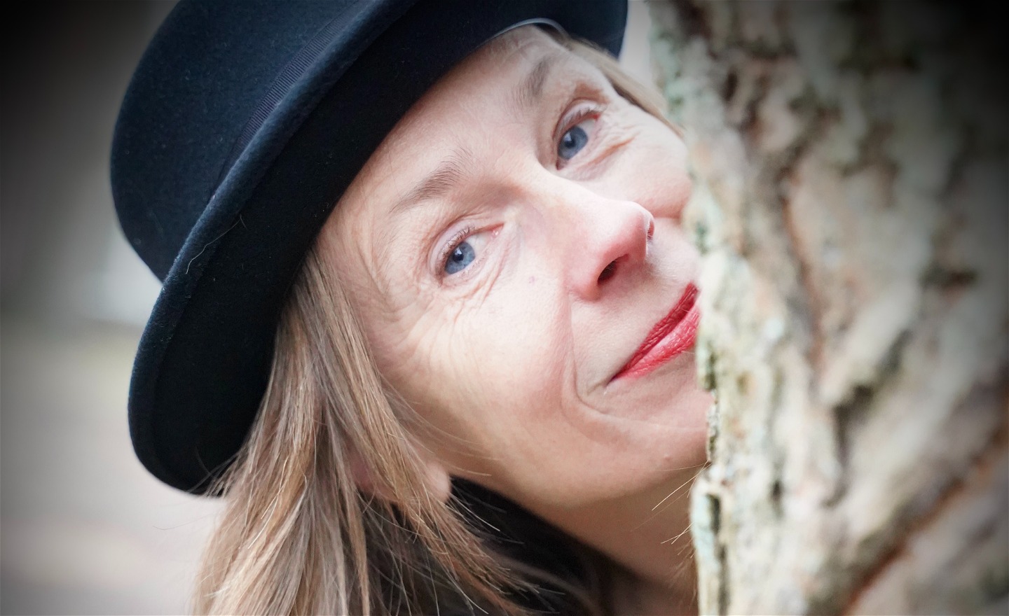 Porträtt på Micaela med hatt som tittar fram bakom en trädstam