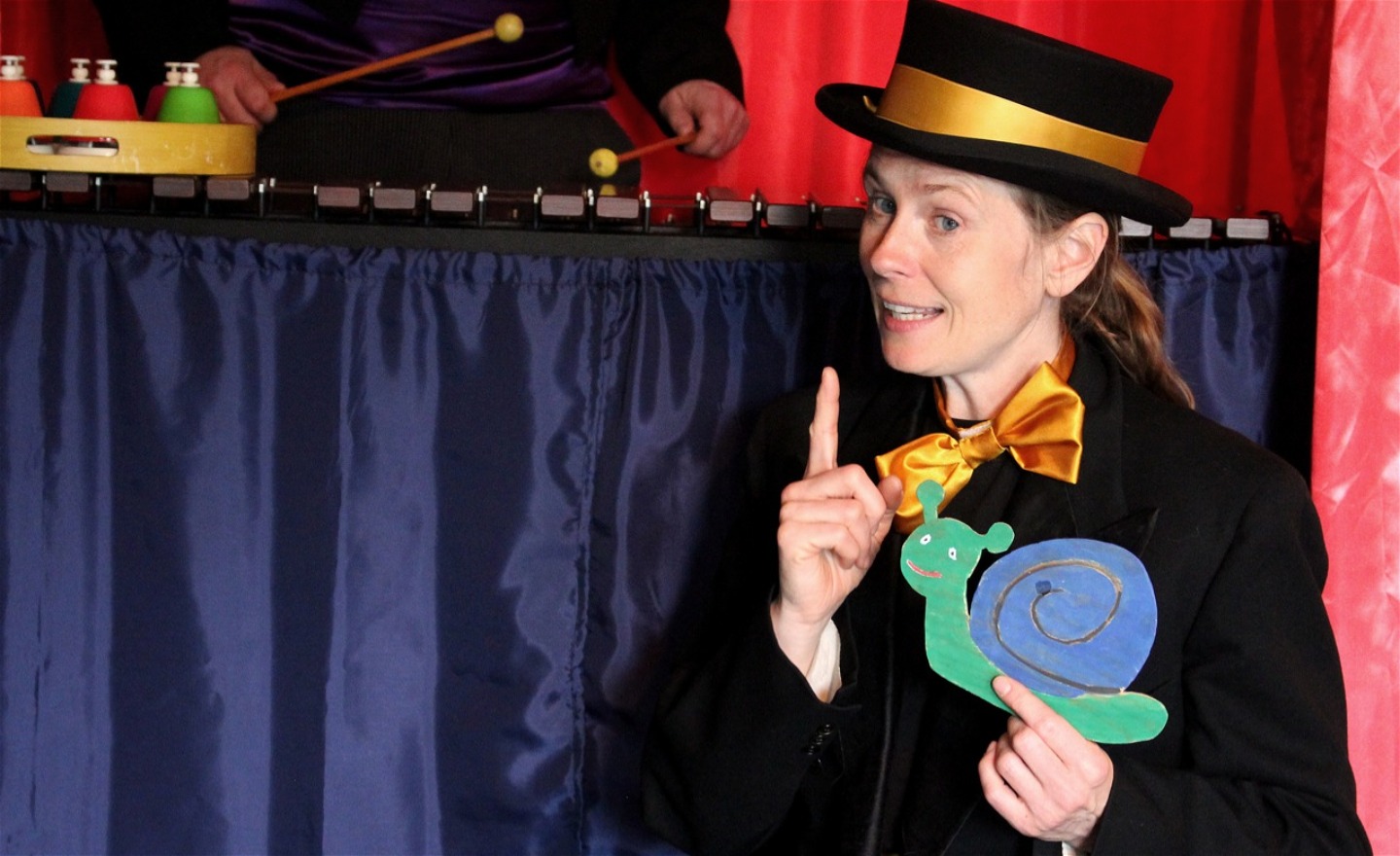 Micaela i höghatt, håller i en snigel, visar pekfingret och sjunger Lilla snigel. I bakgrunden syns Mikaels hander spela xylofon samt klockspel