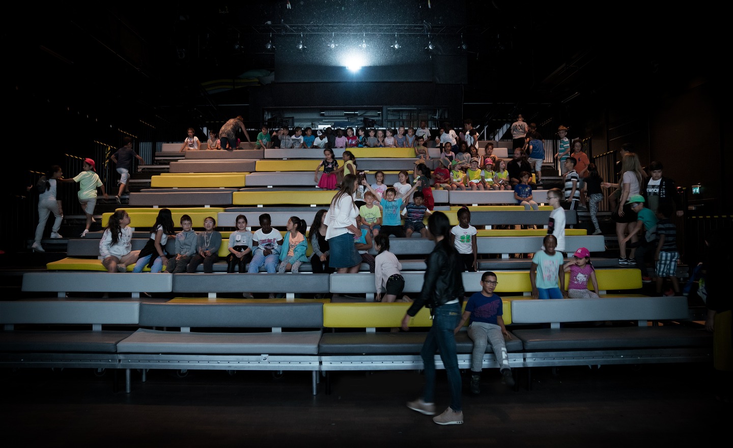 Barn och vuxna sitter och rör sig runt på en stor gradäng med färgglada bänkrader i en mörk salong.