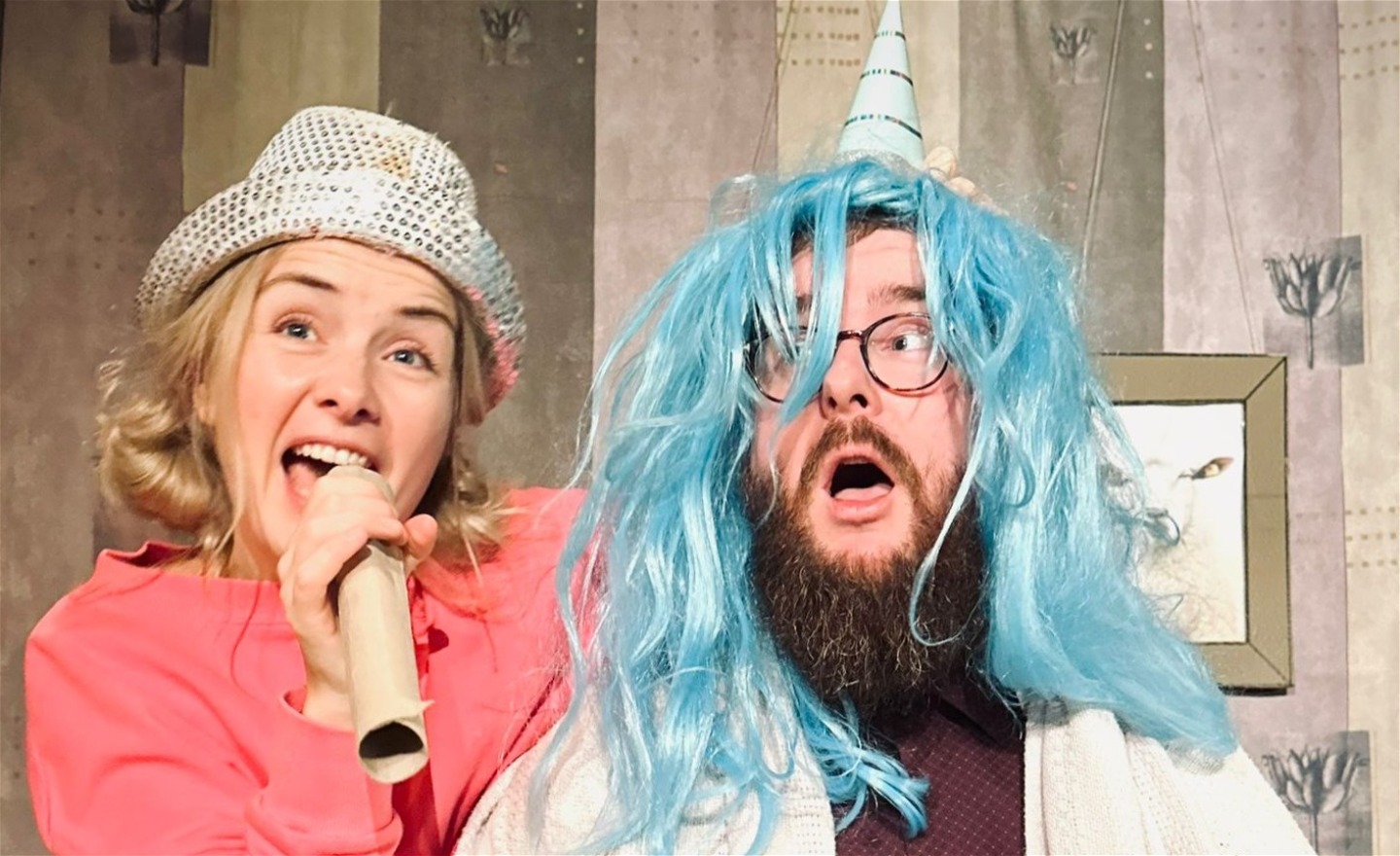 En flicka sjunger i en pappersrulle tillsammans med en man i blå peruk och partyhatt. 