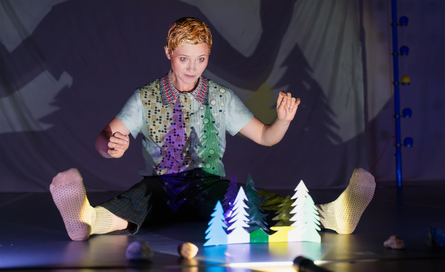 En dansare sitter på golvet i ett mörkt och mystiskt scenrum. Framför henne på golvet står små trädsilhuetter av papper i olika färger. Hon tittar på de små träden, samtidigt som en lampa lyser på dem så att de kastar stora skuggor.