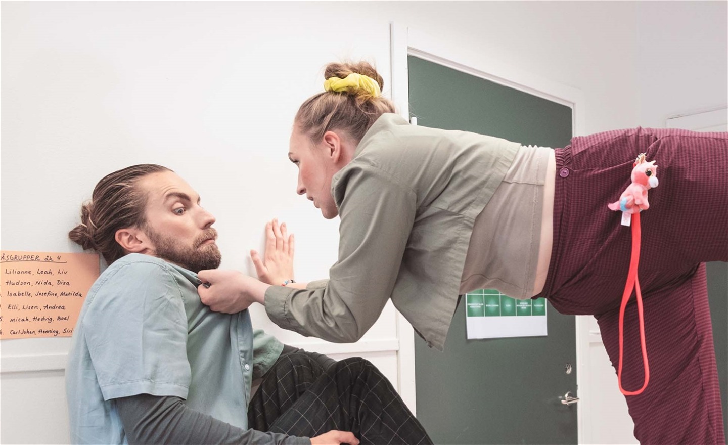 Per sitter uppkrupen på ett skåp i ett klassrum. Katja står på en stol, håller i hans skjortkrage och ser ut att ställa honom till svars för något.