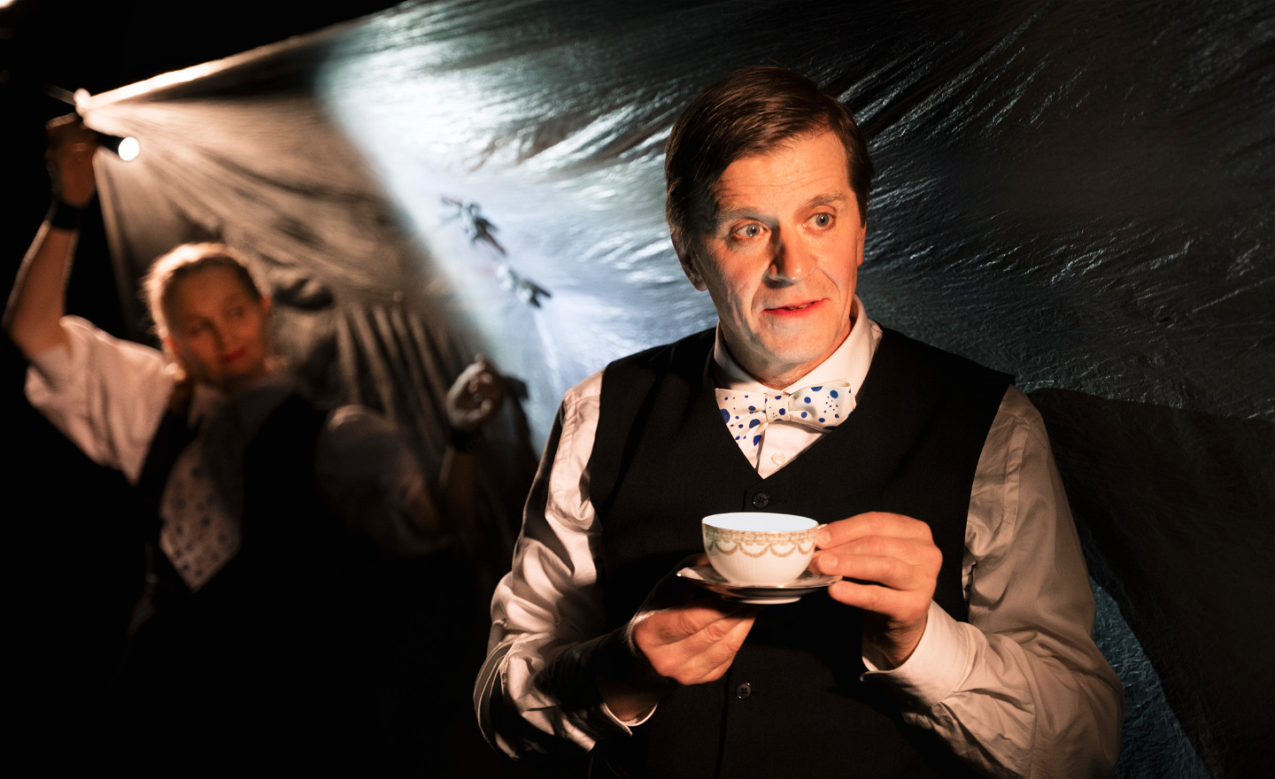 Skådespelare med en kaffekopp. Bakom honom en skådespelare som lyser med ficklampa.