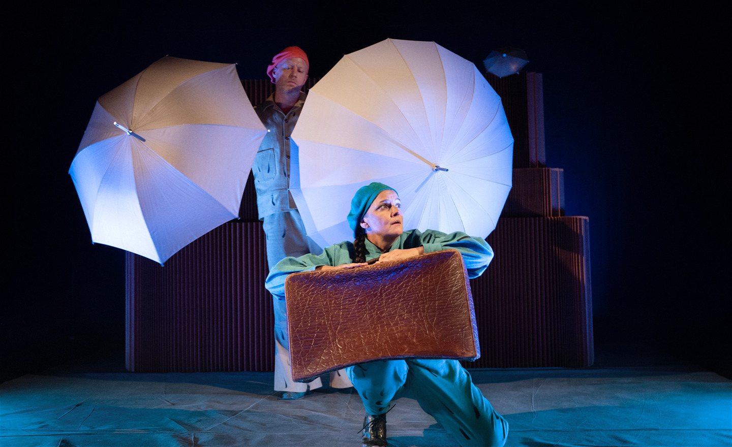 Skådespelare med resväska. Bakom henne en skådespelare med två vita paraplyer