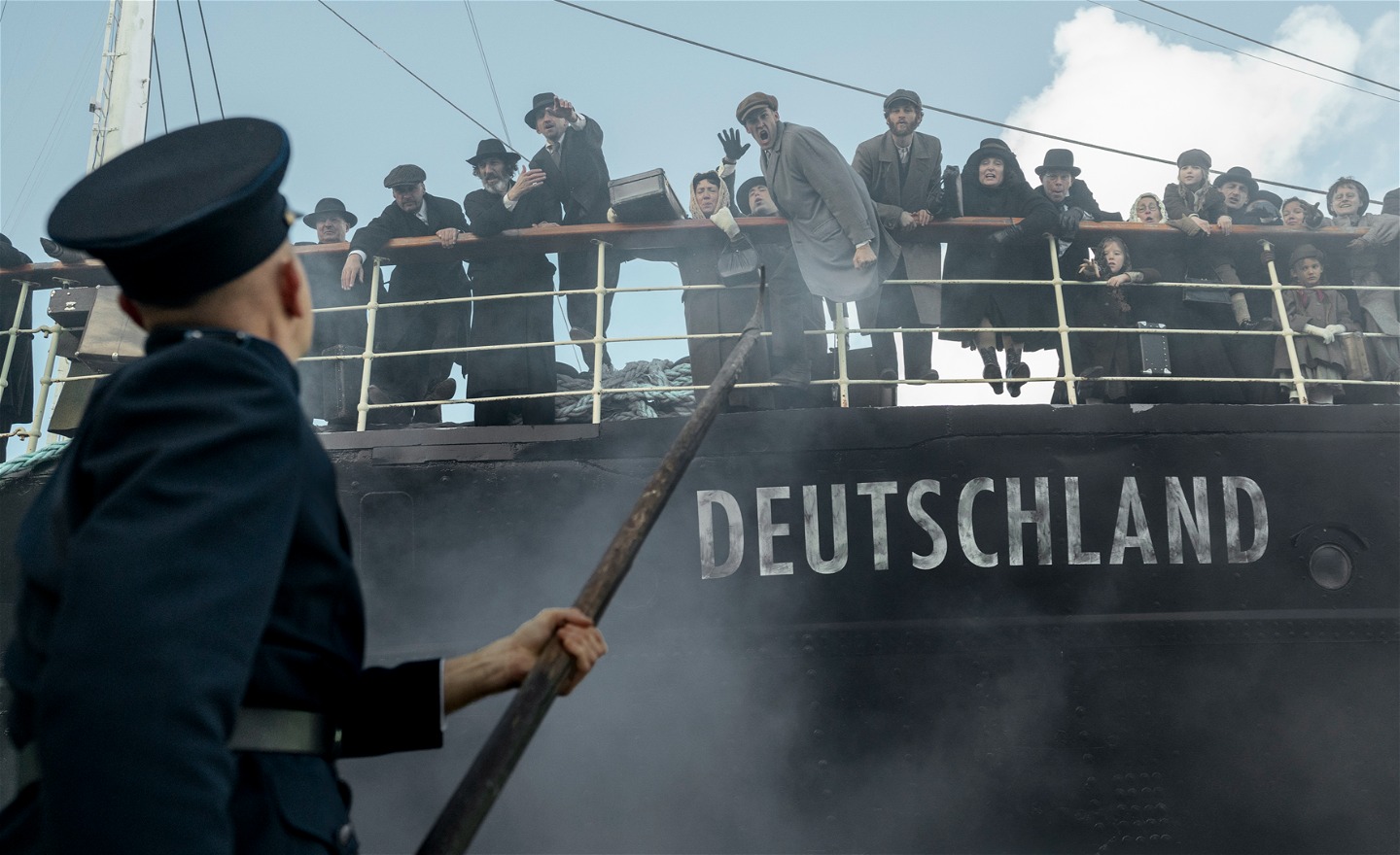 Ett svart fartyg med texten Deutschland på skrovet. Ombord står många människor vid relingen och ropar åt en uniformerad man som står på kajen och riktar en lång båtshake mot fartyget.