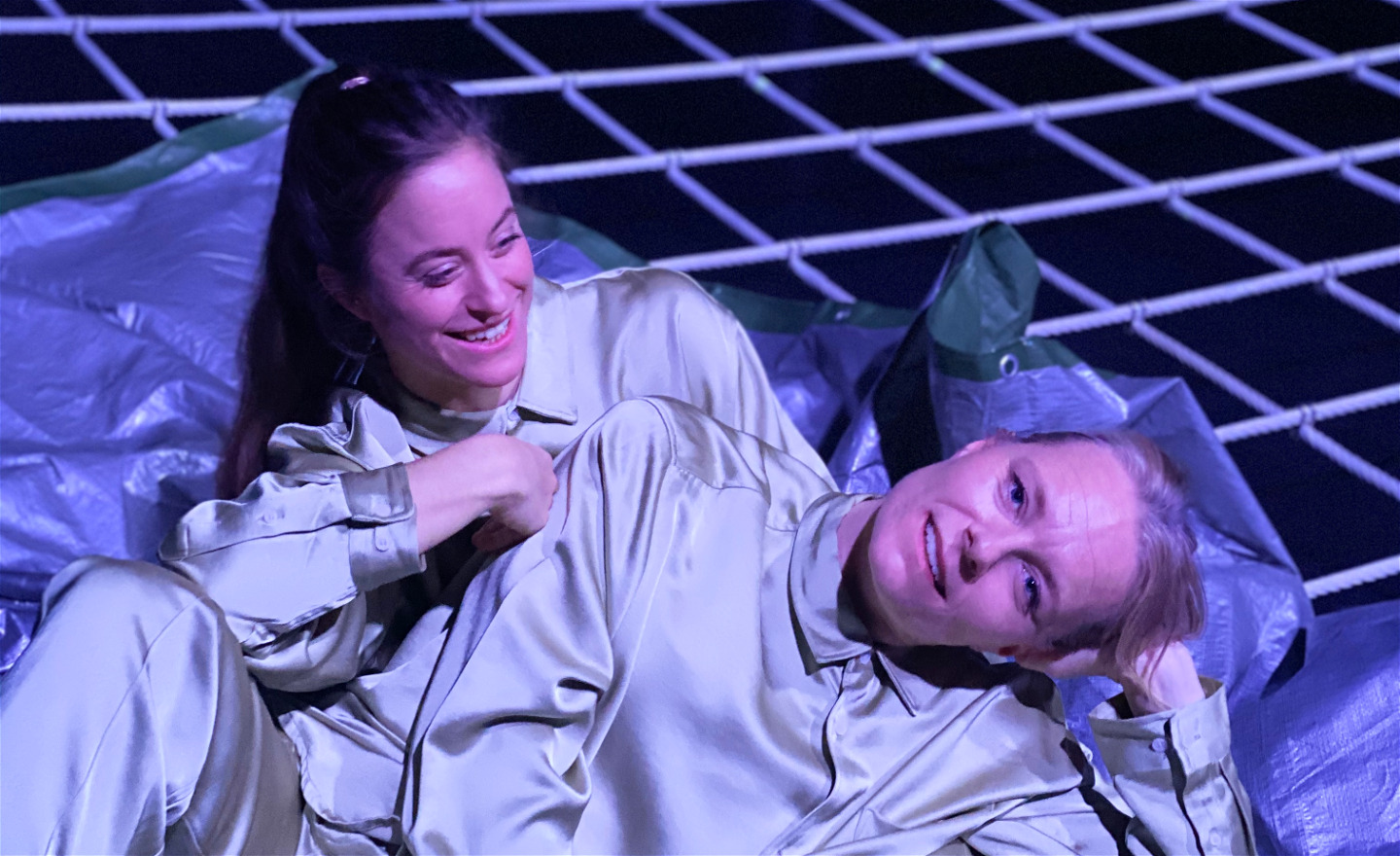 Porträtt på koreograferna Anja Arnquist och Madeleine Lindh. De har på sig beiga sidenkostymer och sitter på en silvrig pressenning, som ligger på ett klätternät, som är en del av scenografin till duons föreställning WEB.