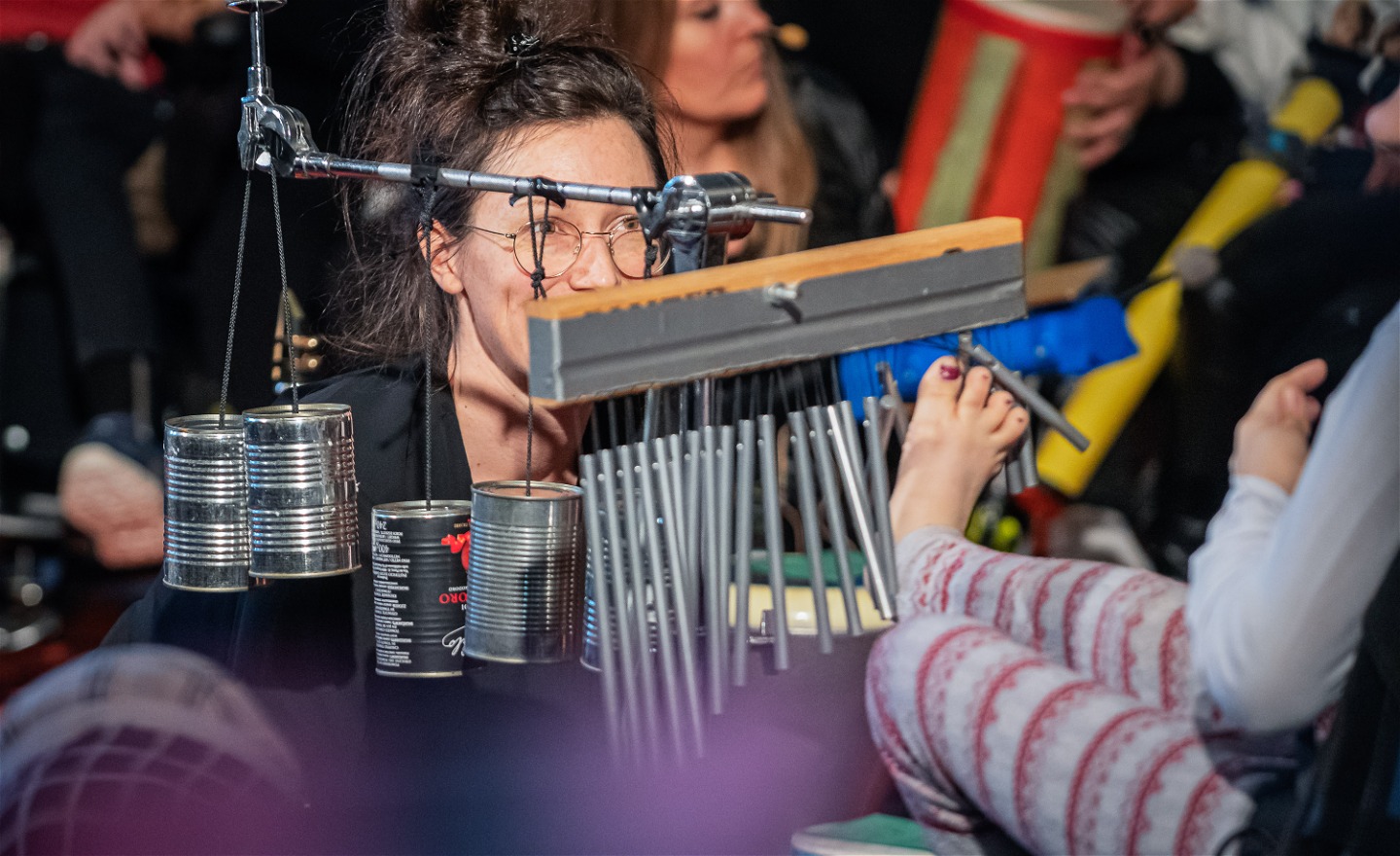 Bilden visar en elev som spelar wind chimes med sin fot. I bakgrunden står Lucy Rugman och Emma Nordenstam och assisterar eleverna med olika slagverksinstrument.