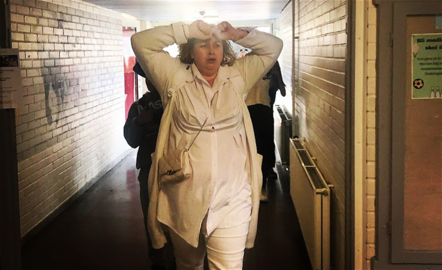 En kvinnlig skådespelare går i en skolkorridor. Ledsen med händerna upp, knytna vid pannan. Bakom elever.