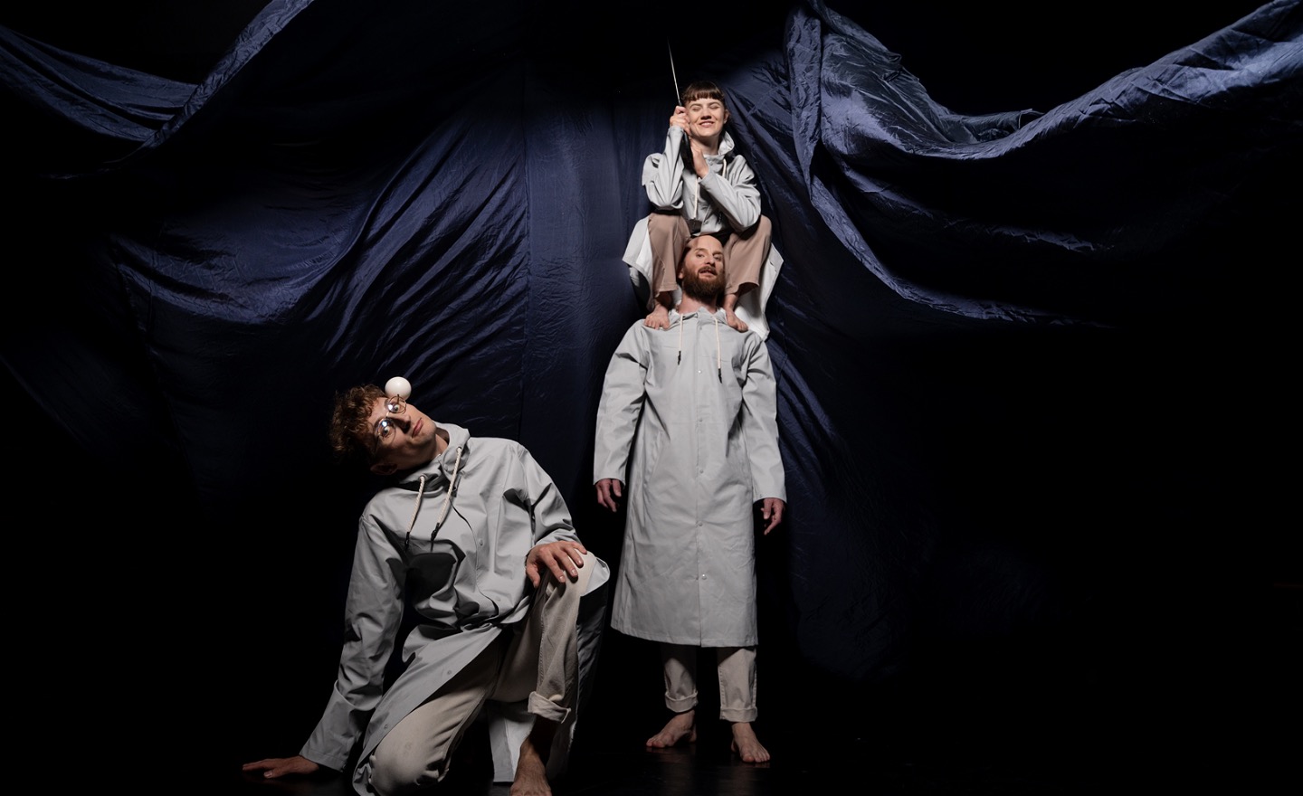 Tre artister i grå regnrockar står framför ett blått, böljande tyg. En sitter på en annans axlar och håller i ett paraply. En annan står bredvid och balanserar  en jongleringsboll på sidan av huvudet.