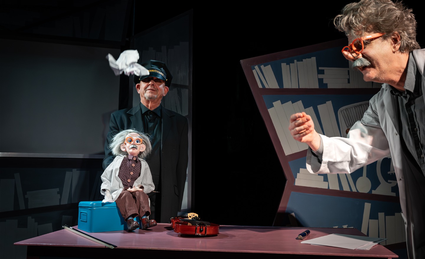 Einstein försöker att förklara tyngdkraften genom att kasta upp hopknycklade papper, som förstås genast faller ner. Dockan Albert med skötare Lazy tittar på.