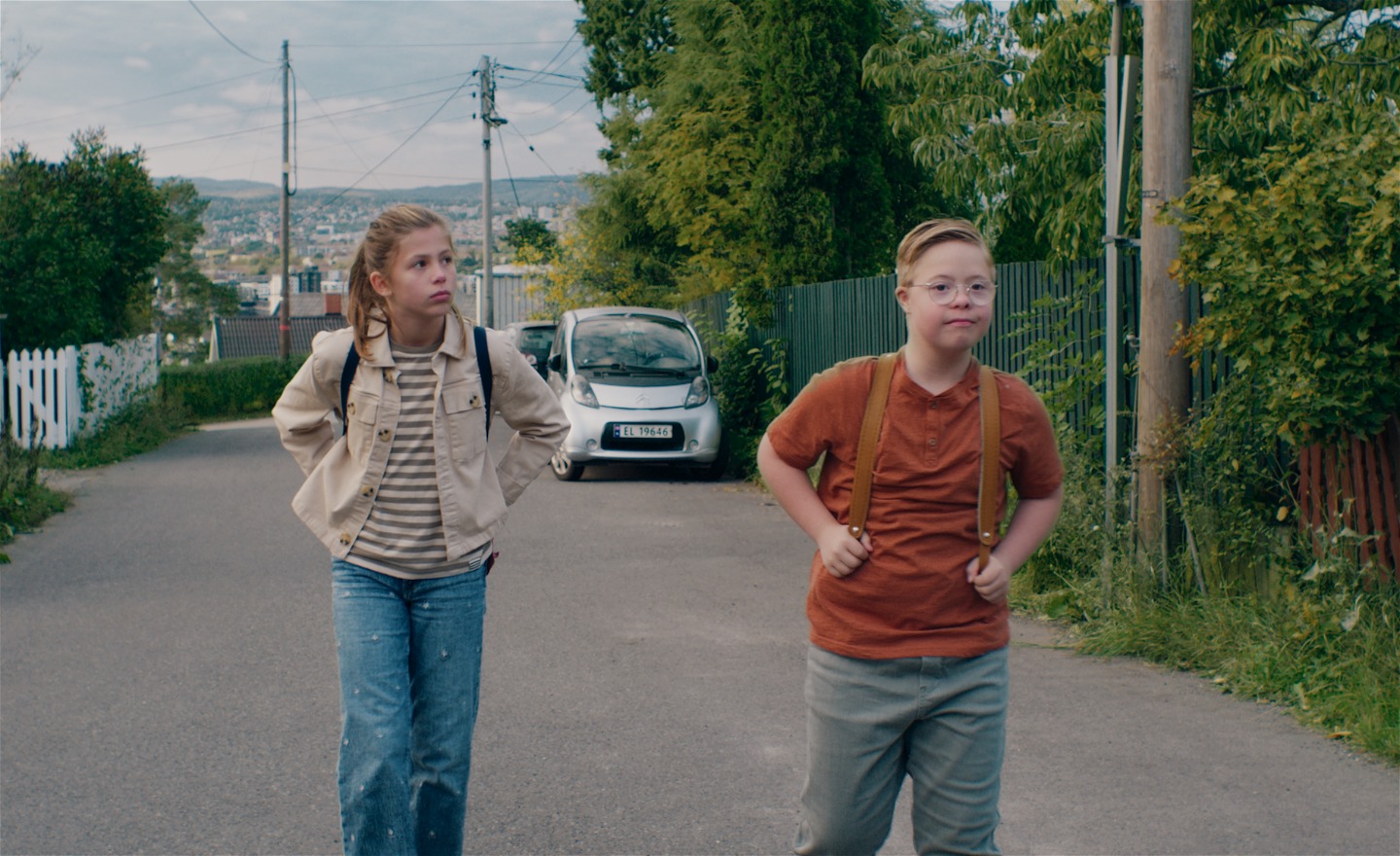 En flicka i mellanstadieåldern går på en asfalterad gata, snett bakom en pojke med downs syndrom. Båda ser allvarliga ut.