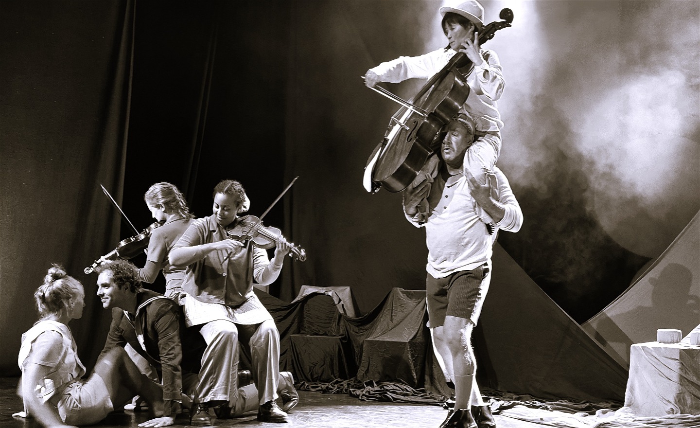 Två violinister, en sångare och en ung kvinna sitter på scengolvet och på stolar. En man bär en cellist på sina axlar samtidigt som hon spelar Händels musik.