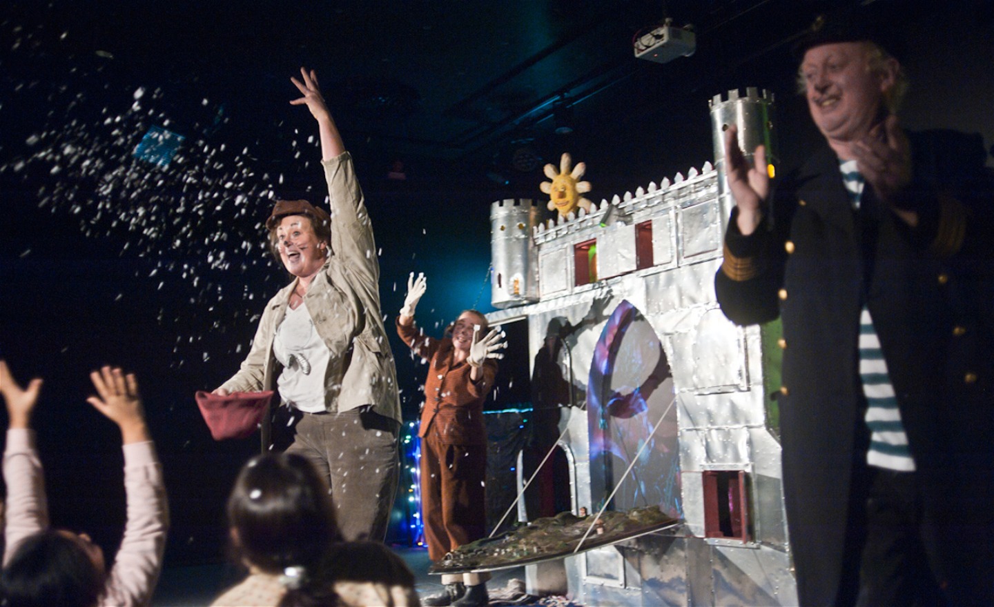 Tre skådespelare står på en scen med ett silverfärgad slott i bakgrunden.