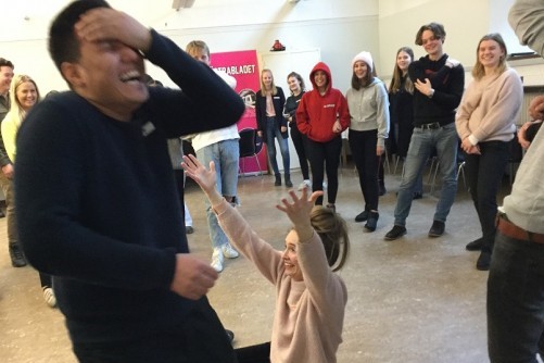 En person tar sig för pannan och skrattar, en person sitter nedanför och lyfter på armarna och en hel grupp står runt om och tittar på.