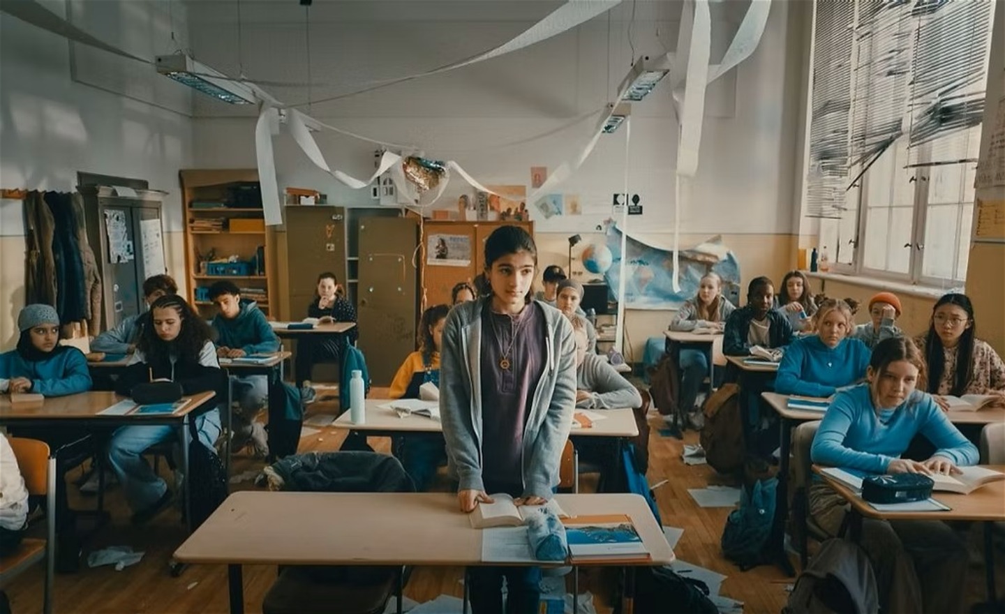 En tonårsflicka står vid en bänk i ett mycket stökigt klassrum. Framför henne ligger en uppslagen bok.