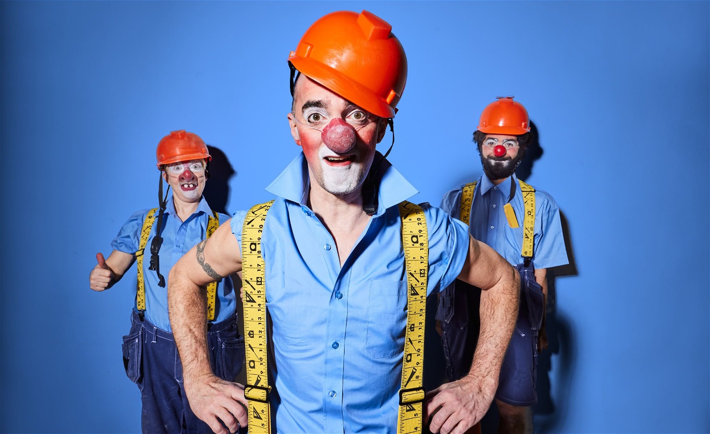 Tre clowner i byggarbetarkläder, orange hjälmar,  blå skjortor och blåa byxor. 