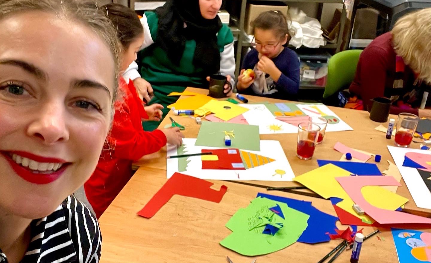 Foto föreställande närbild på Hanna som ler mot kameran. I bakgrunden syns barn och vuxna och ett bord med färgglada papper och papperscollage.