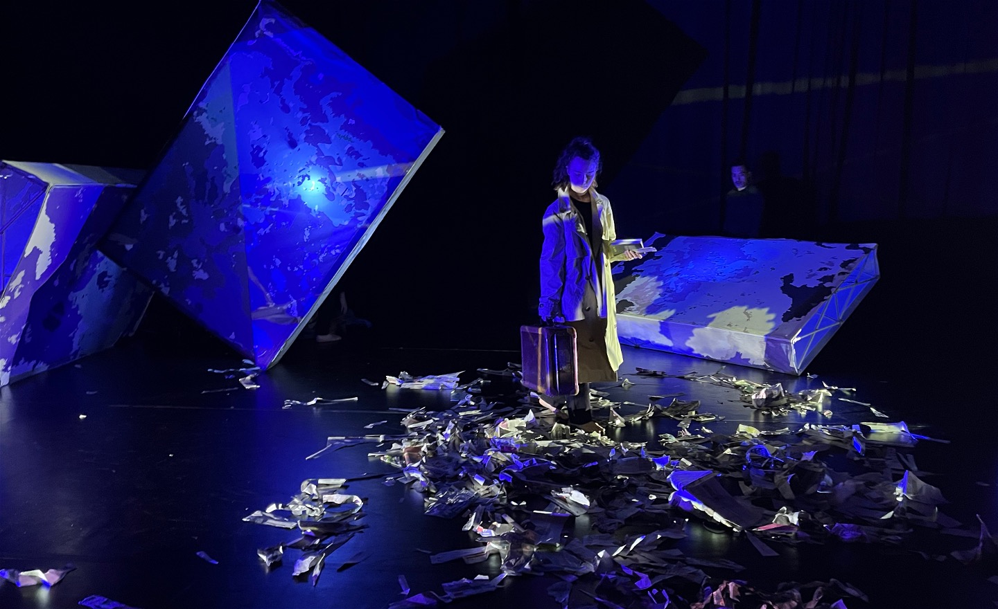 Lutande skärmar, sönderrivna tidningar på golvet, en dansare med resväska går över scenen