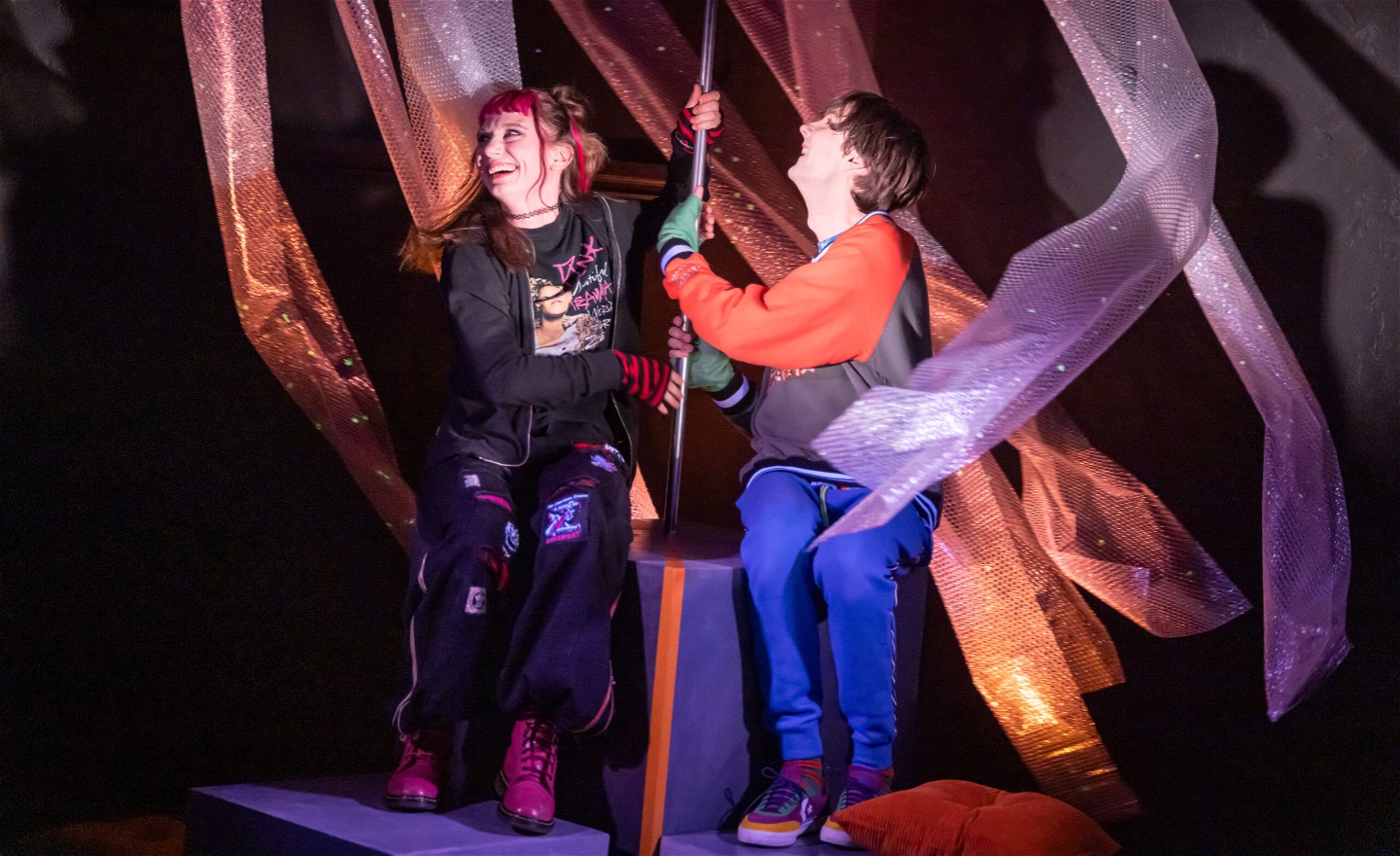 Bild från föreställningen. En tjej och en kille sitter och skrattar och håller i en pinne som är fäst i en låda och runt omkring hänger remsor som rör sig runt dem. 