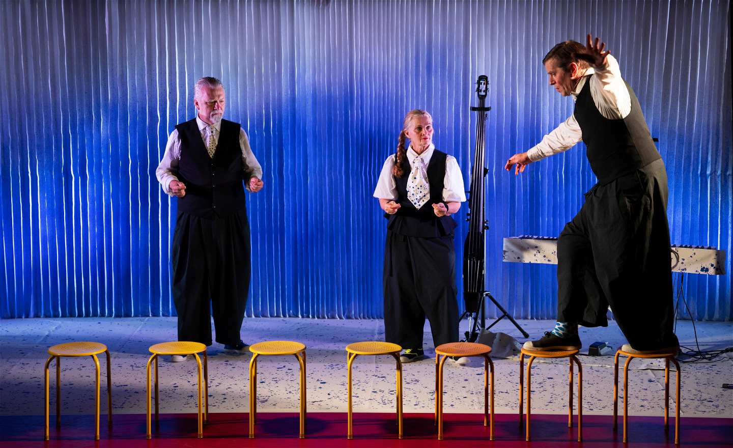 Två skådespelare betraktar en tredje som balanserar på gula pallar.