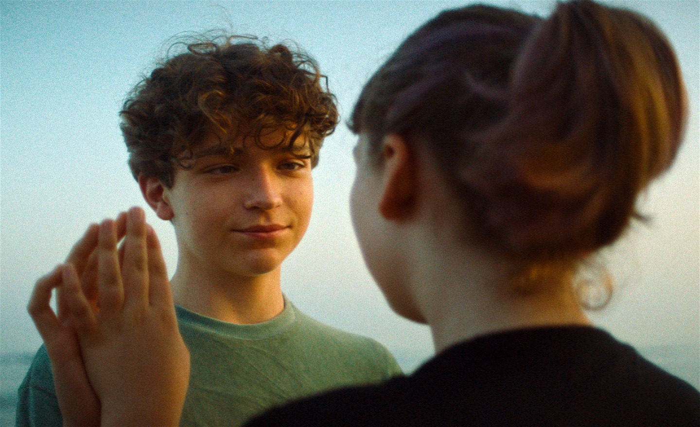 Blå himmel i bakgrunden. En pojke och en flicka står och ser på varandra. Deras händer rör vid varandra.