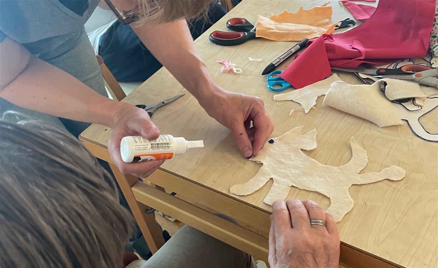 Ett bord med materail som textil och saxar. Två personer hjälps åt att göra en tyghund som lyfter på benet för att kissa.