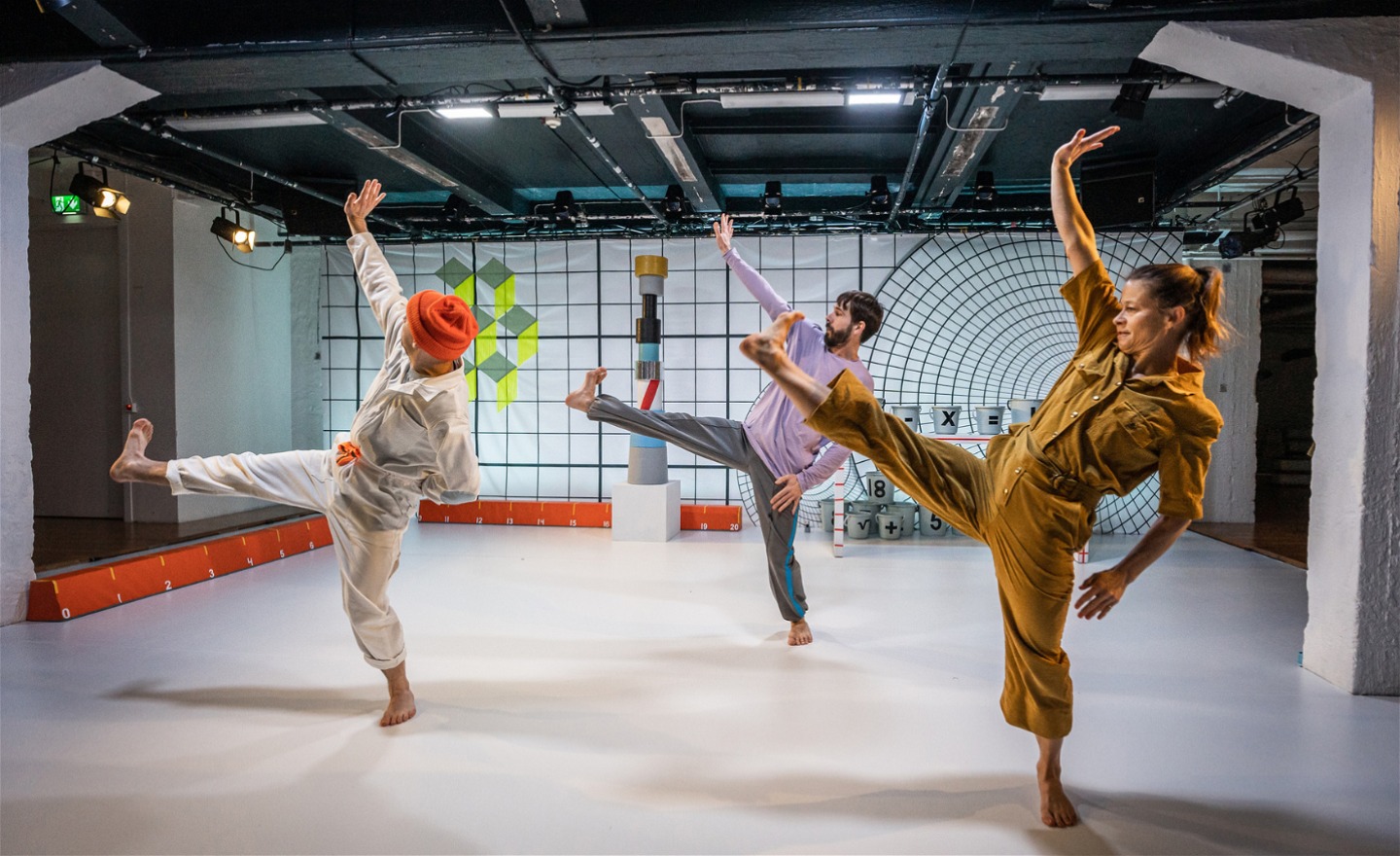 Tre dansare som sträcker sitt högra ben och sin högra arm uppåt. Färggranna figurer i bakgrunden. 