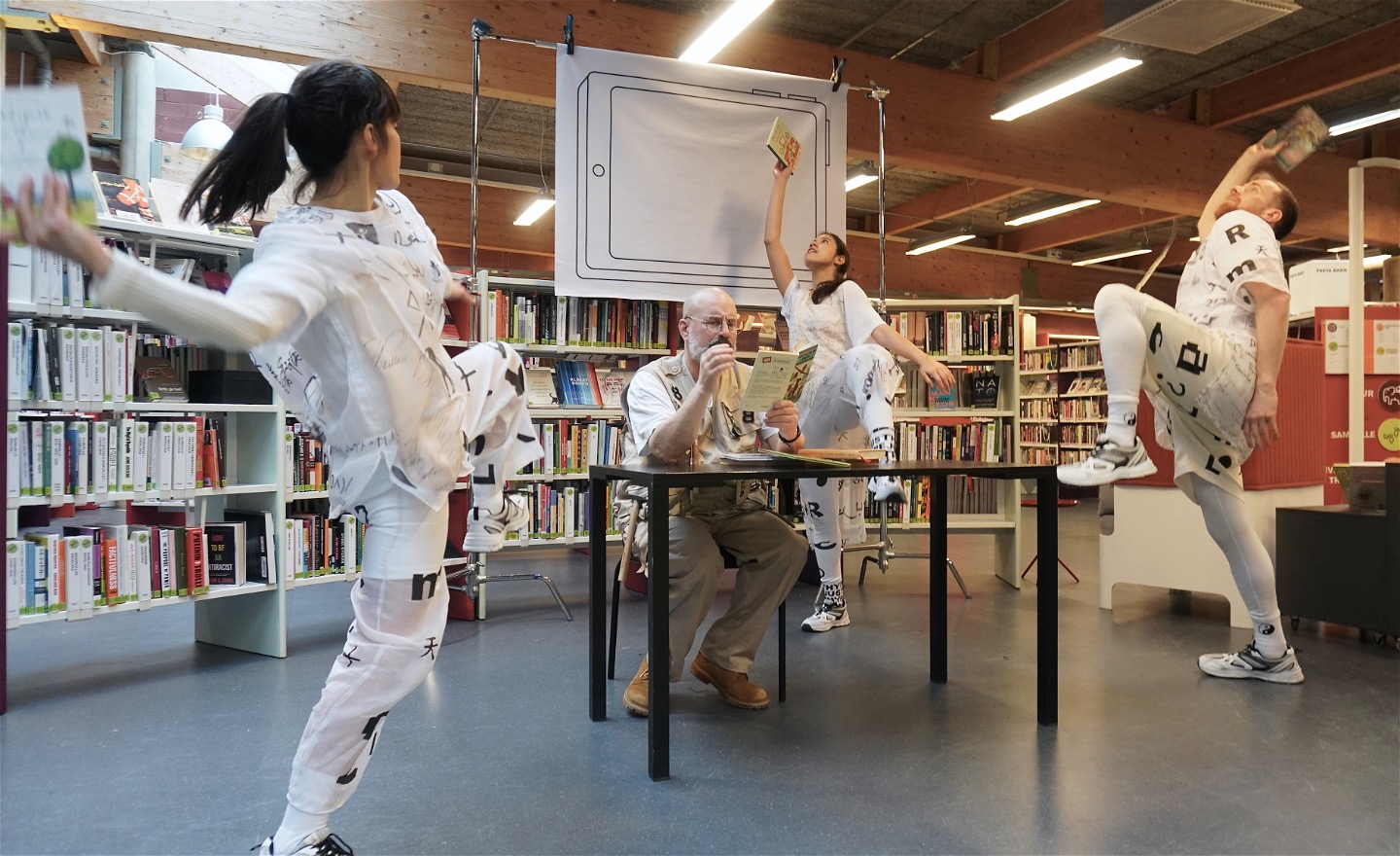 3 dansare med varsin bok i handen lutar sig bakåt i en energifylld rörelse samtidigt som de lyfter på ena benet, en skådespelare sitter vid ett bord i mitten och läser en bok