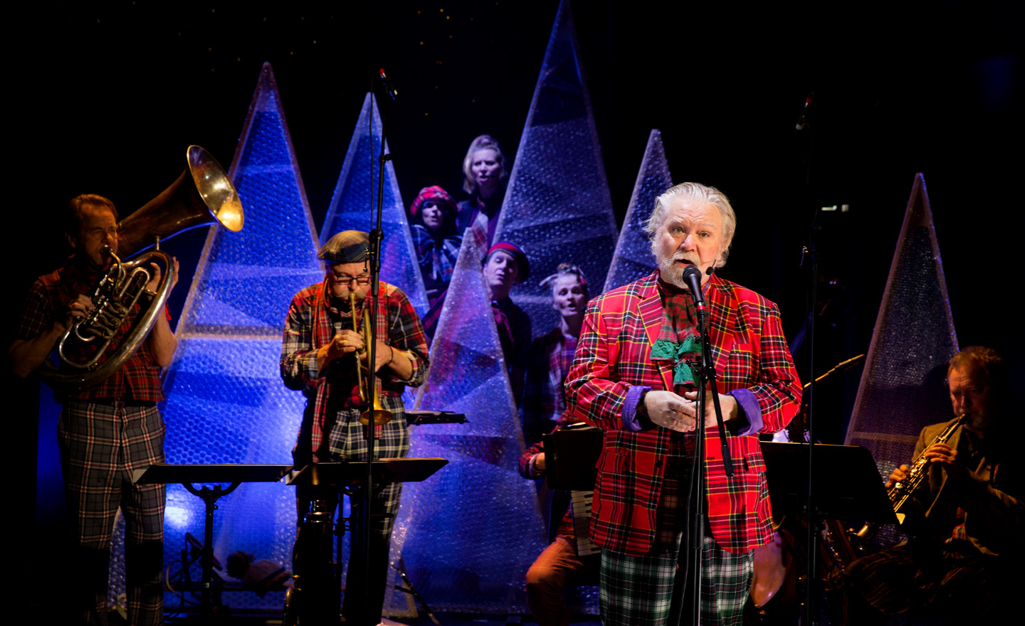 Skådespelare i rödrutig kostym vid mikrofon tillsammans med musiker.