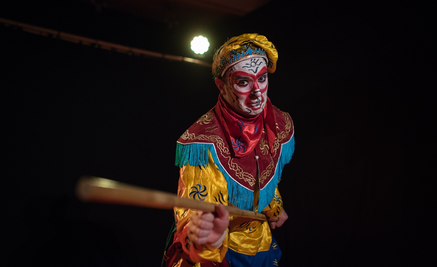En skådespelare sminkad till apa i ansiktet och klädd i traditionell kinesisk teaterkostym tittar in i kameran, hållandes en trästav i handen.