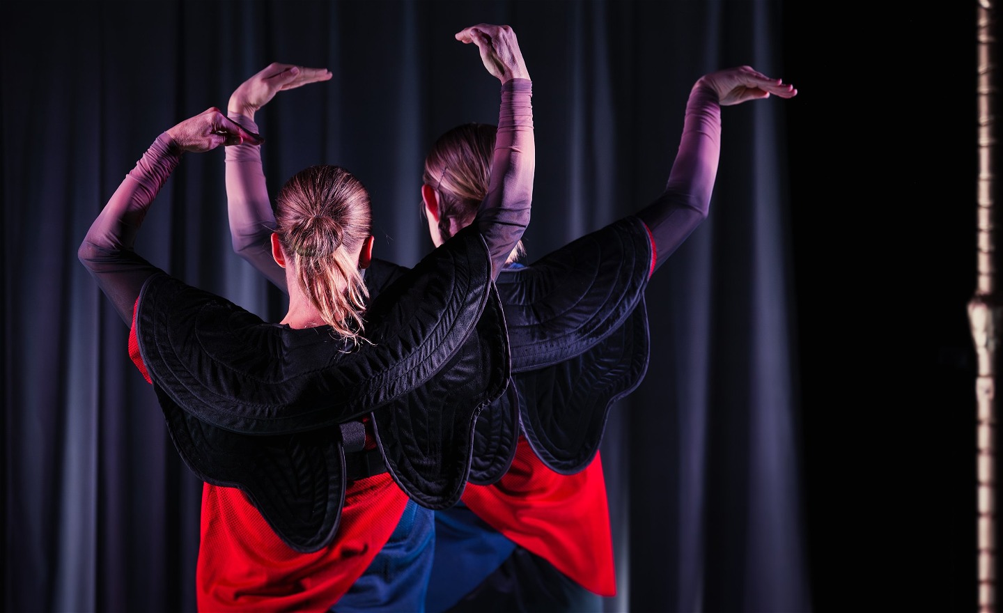 Två personer står med ryggarna mot kameran med händerna som näbbar i luften ovanför huvudet.