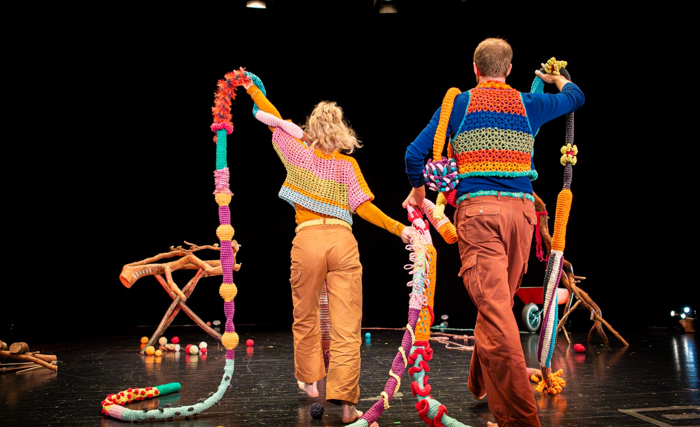 Två personer med ryggen emot oss. Dom bär på långa stickade färgglada band. 