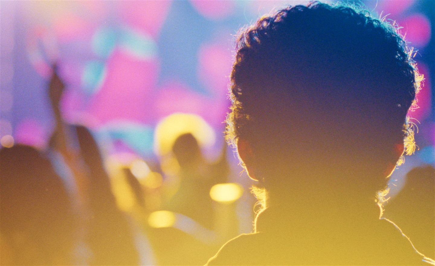 En pojke står med ryggen mot kameran. Framför honom dansande människor och belysning i olika färger i ett varmt ljus.
