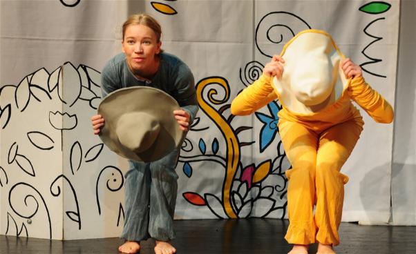 Två dansare i färgglada overaller, en gul och en turkos, gömmer sig bakom hattar.