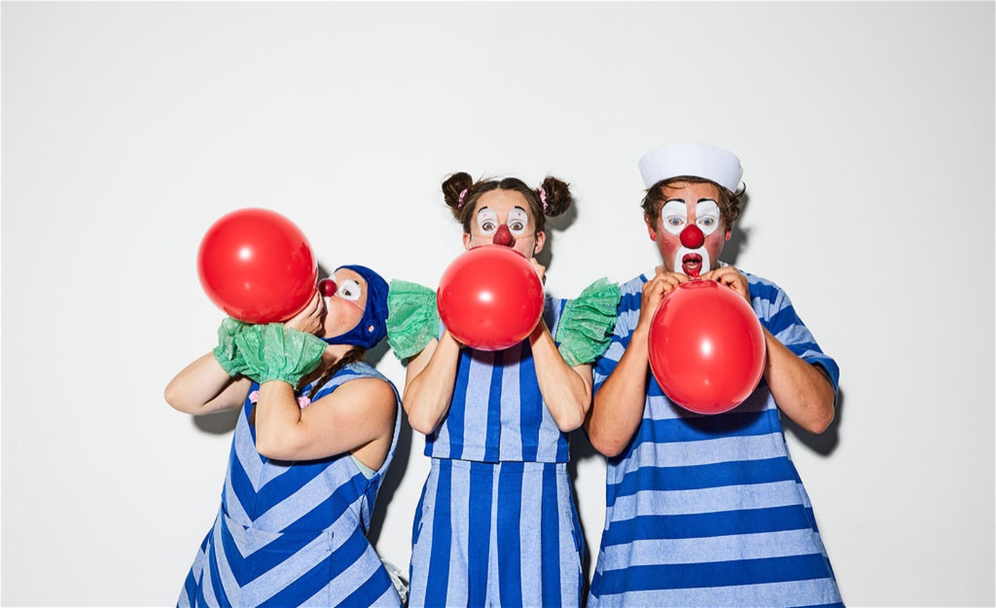 Tre clowner i randiga kläder blåser upp varsin röd ballong.