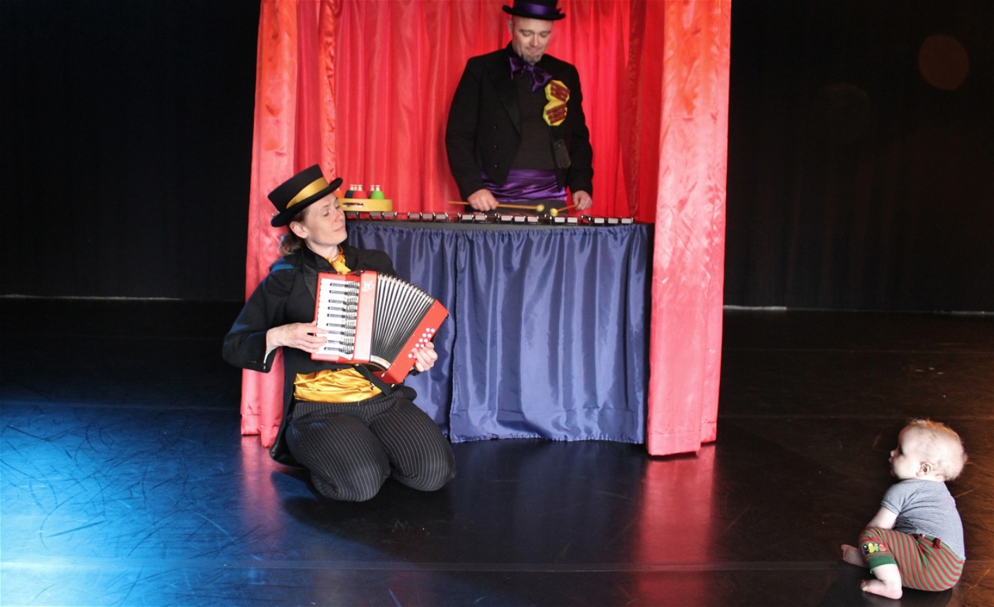 Micaela spelar dragspel och Mikael spelar xylofon för en bebis som sitter framför dem på golvet 