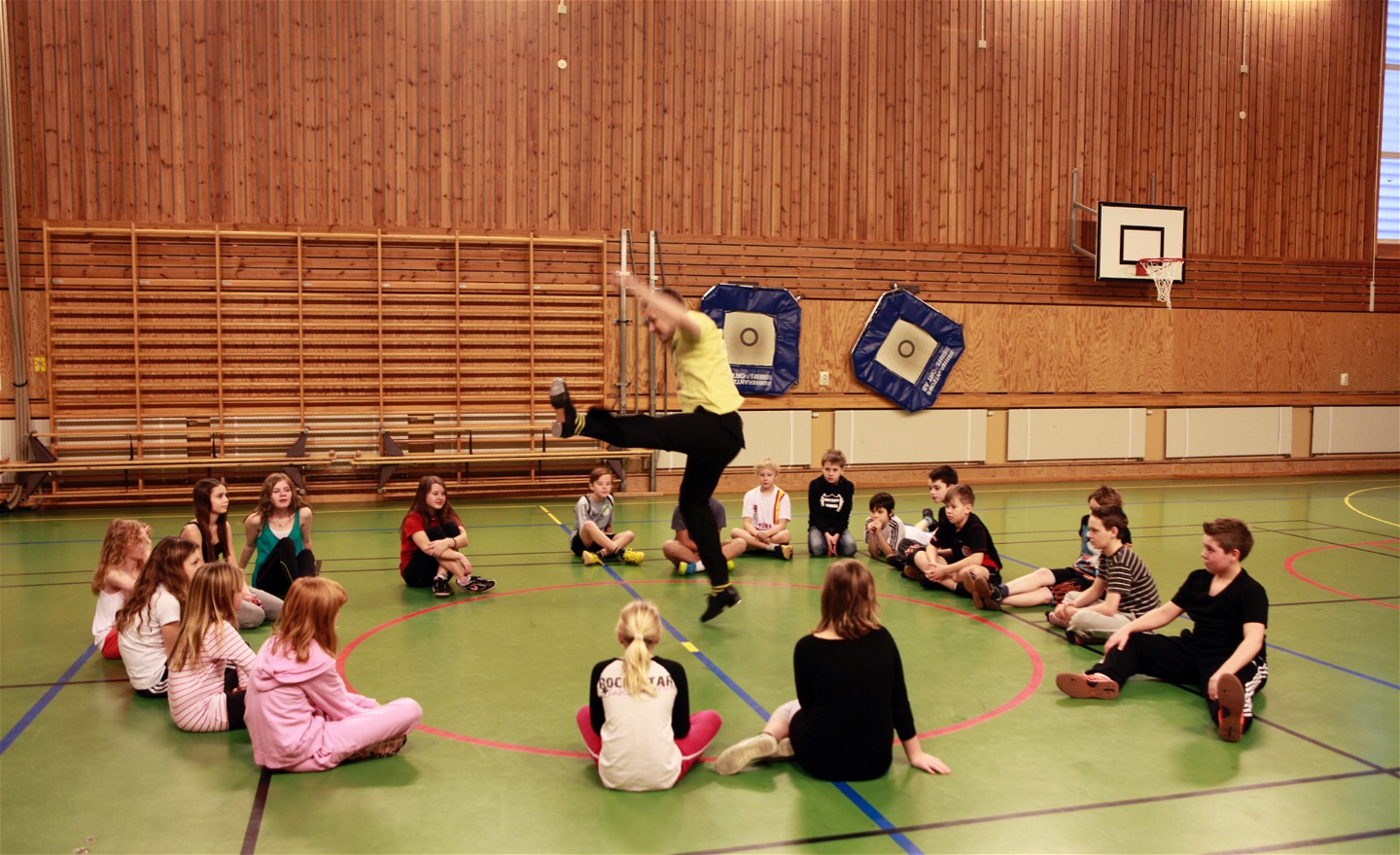 Barn som sitter i en ring runt en dansare med steppskor som hoppar med ena benet högt upp i luften.
