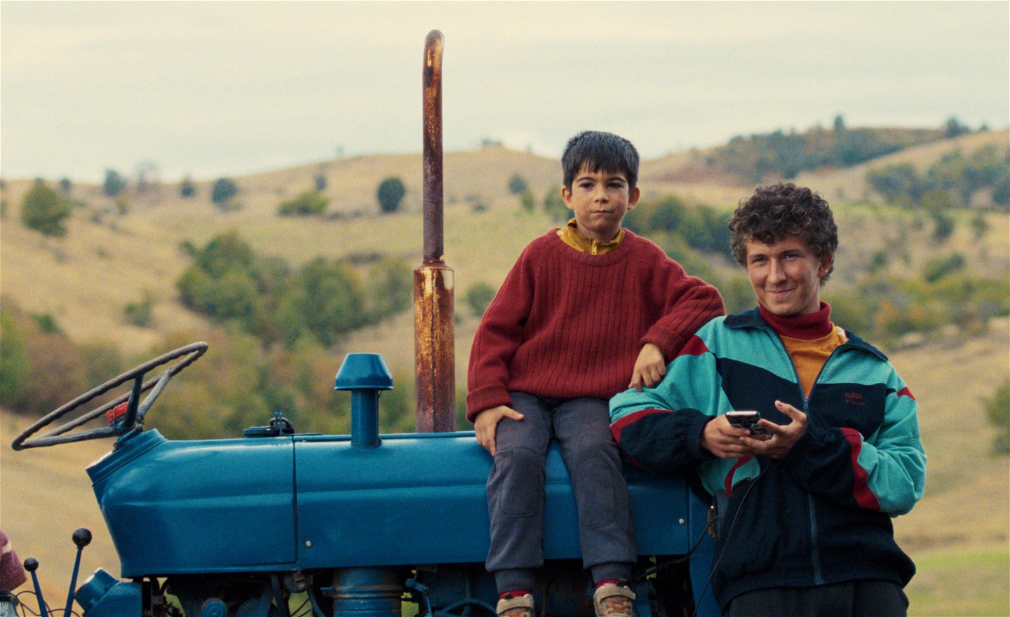En storebror, en lillebror och en traktor. I bakgrunden landsbygd med gröna kullar.