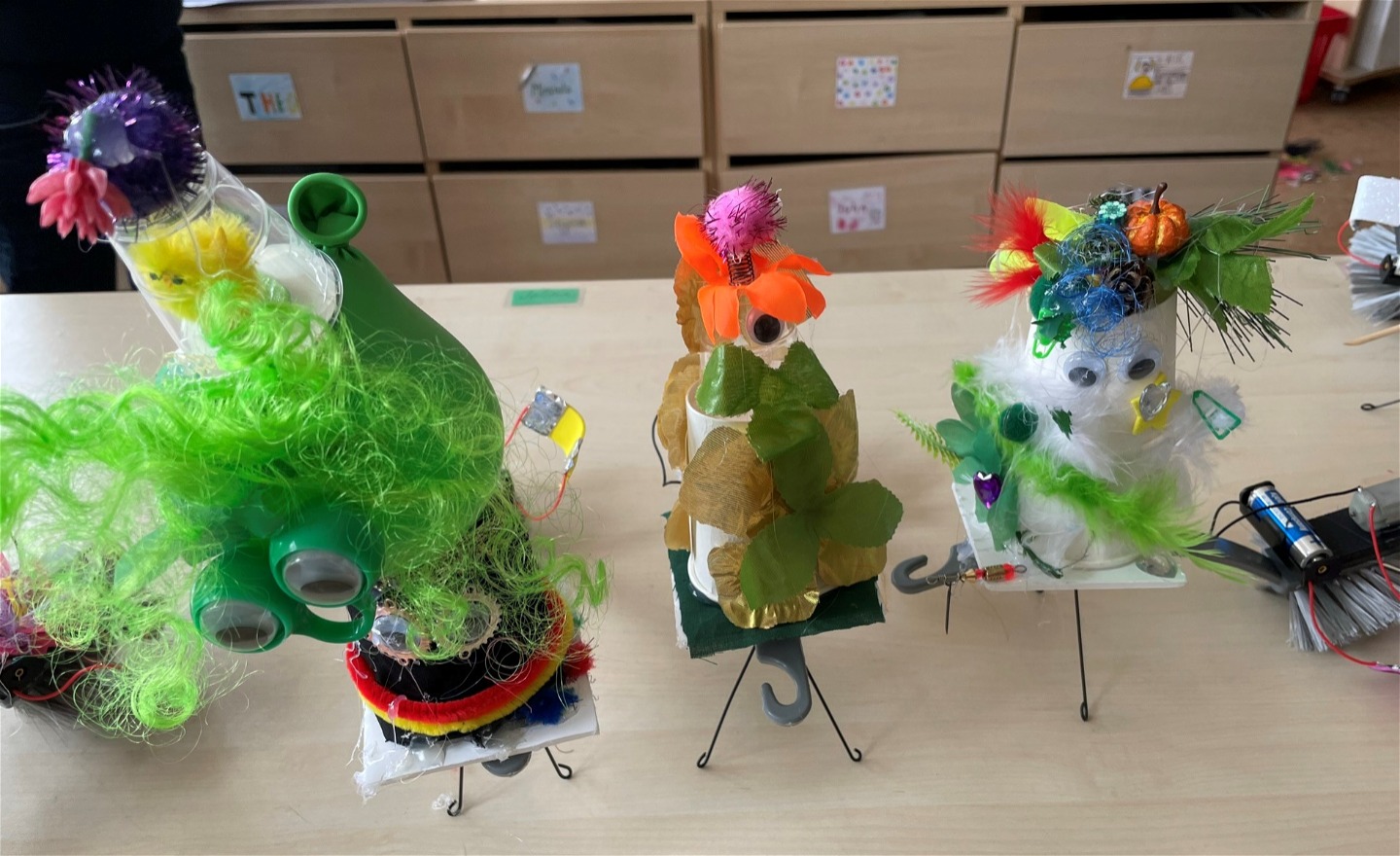 Tre robotar på bordet. En med grön luddklänning En med mycket blad och en blomma och en med blommor i håret
