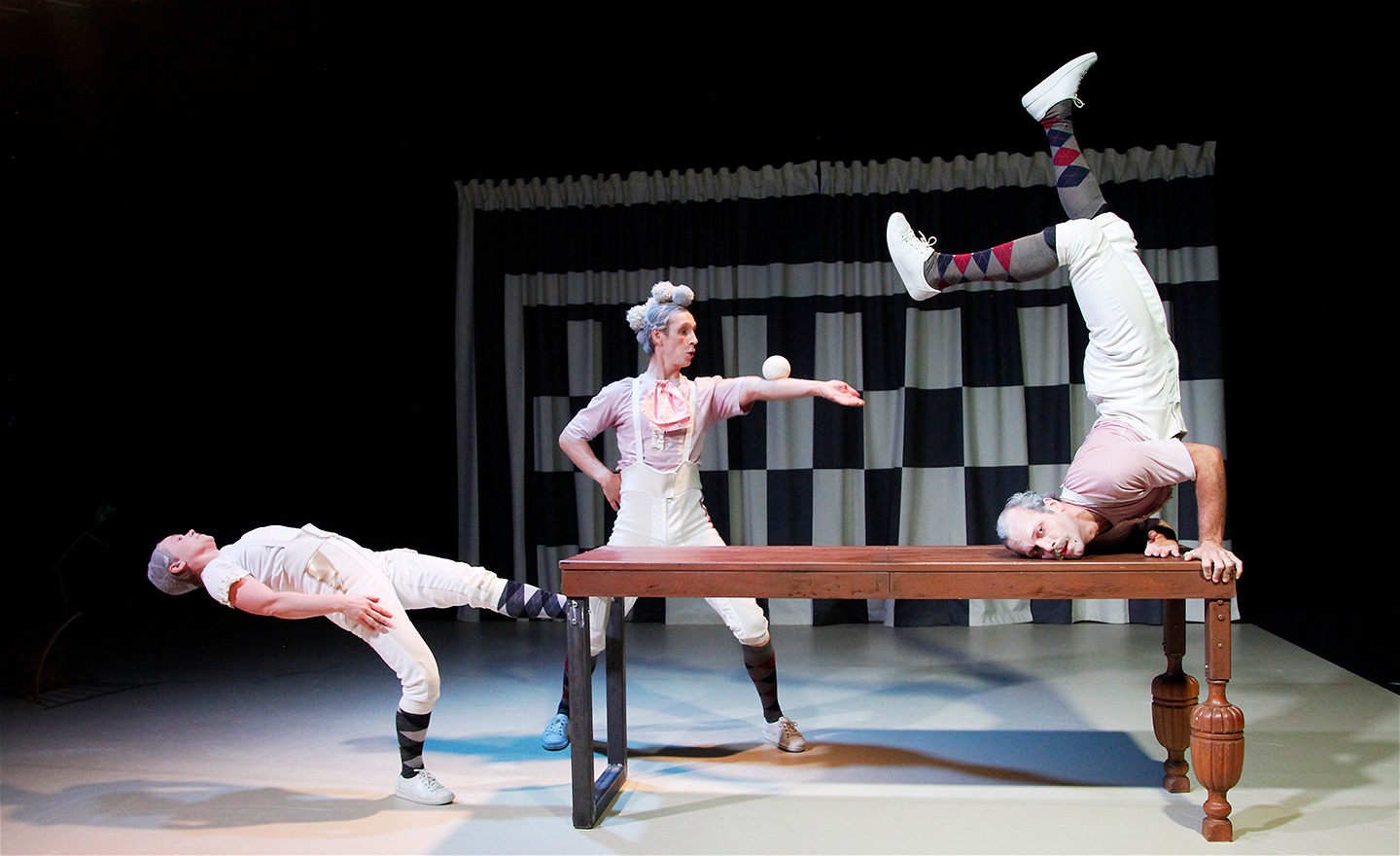 Tre dansare i vita kläder och rokoko-influerat smink och peruk rör sig akrobatiskt kring ett bord: Längst till vänster lutar sig en dansare bakåt i en båge, dansaren i mitten jonglerar med en boll och dansaren längst till höger är uppflugen på bordet